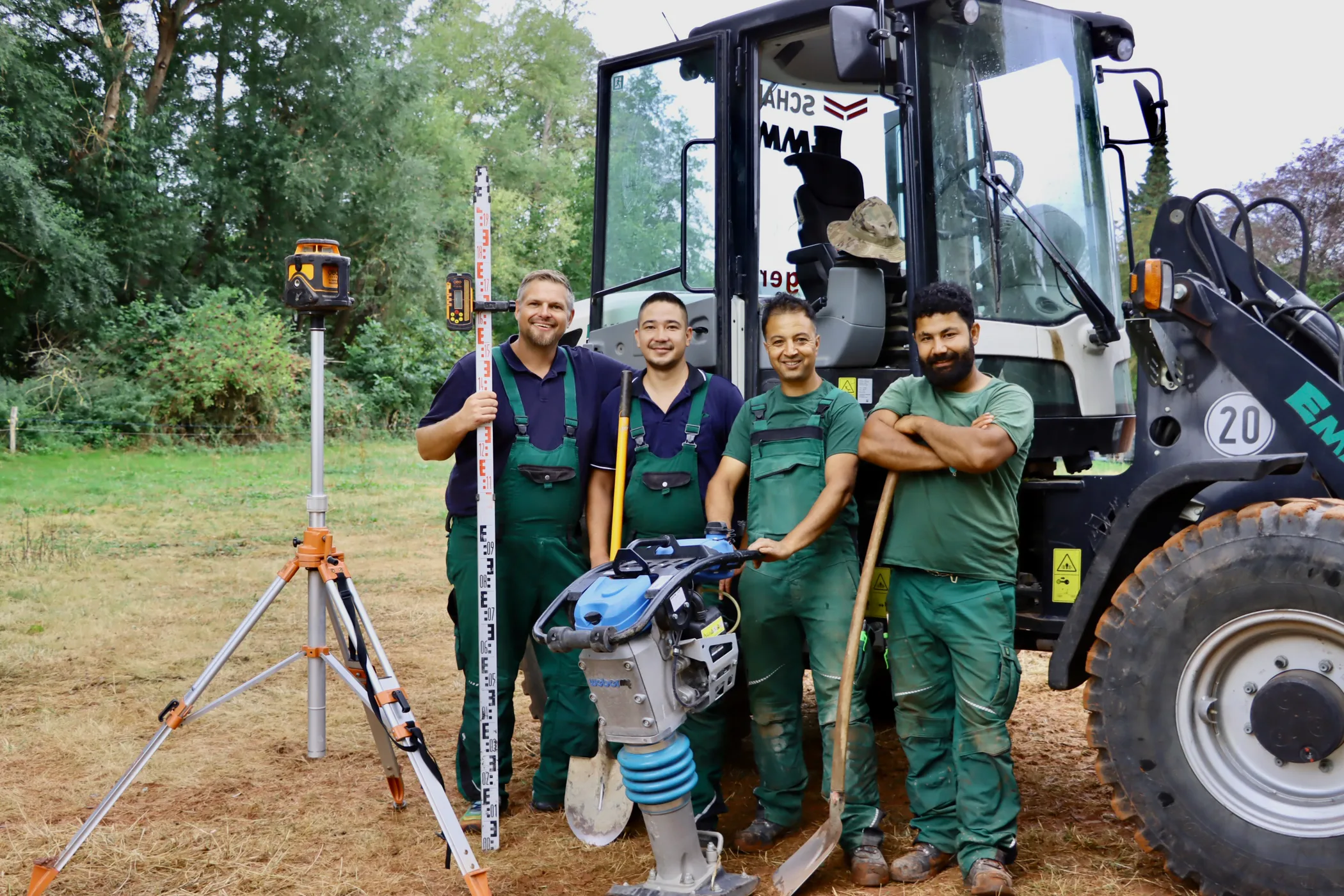 Das Team von Royal Garden mit Rotationslaser, Messlatte und Rüttelplatte vor dem Radlader