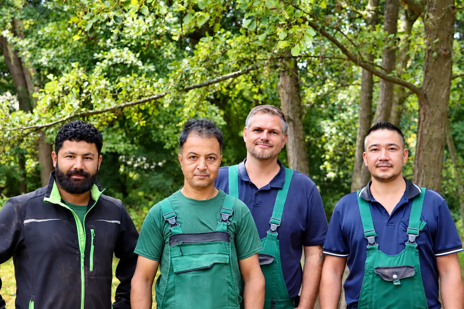 Vier Mitarbeiter von Royal Garden in Arbeitskleidung vor einer alten Baumgruppe